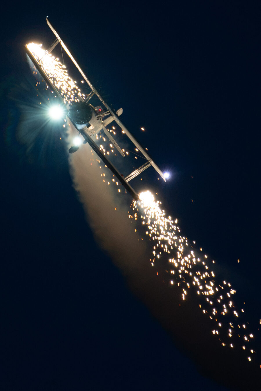 The AgCat flying at night with wingtip pyrotechnics throwing sparks across the dark sky