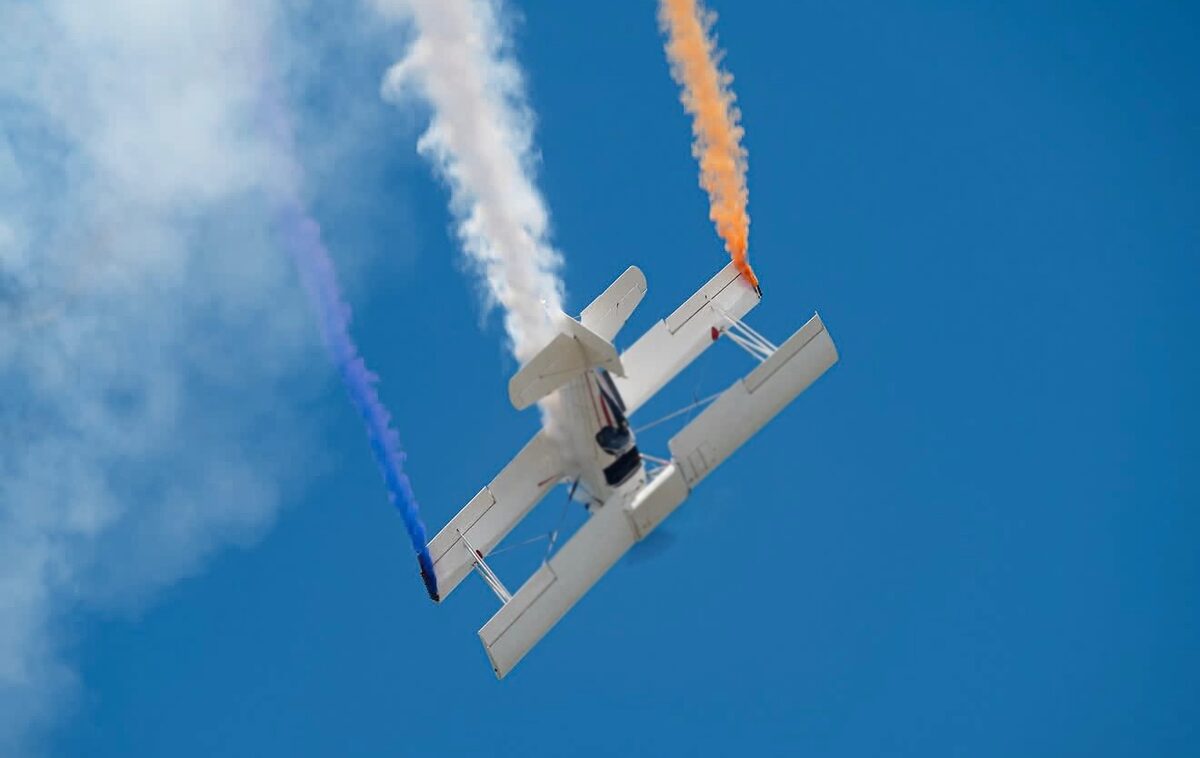 The Grumman G-164A AgCat inverted against a blue sky, trailing blue, white and orange smoke