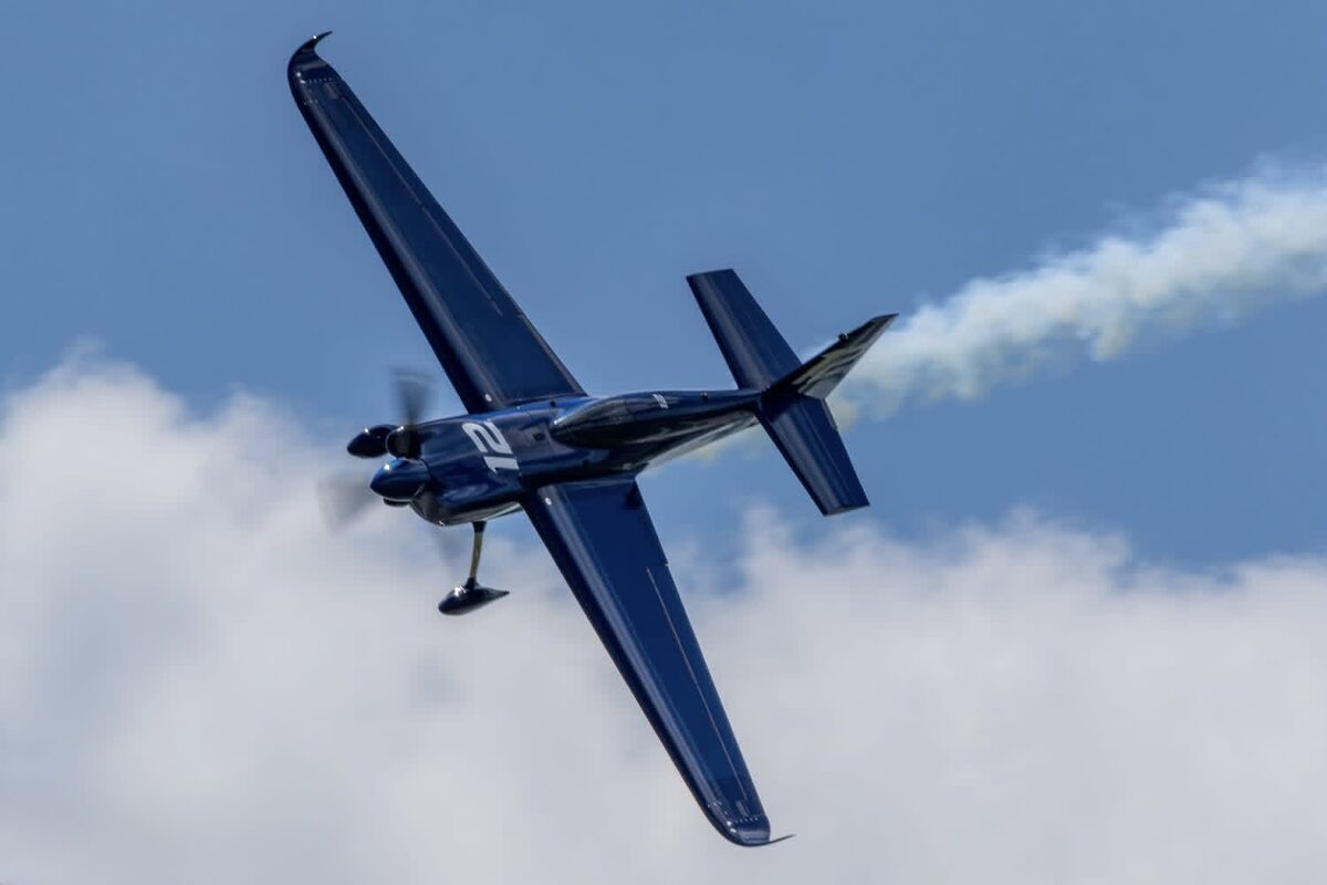 Topside view of the blue Zivko Edge 540 v3 wearing race number 12, banking hard with a smoke trail